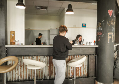 Junge steht vor Bar mit Barhockern, im Hintergrund arbeitet eine Frau und ein Mann